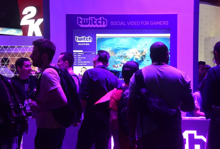 A group of young people standing under purple lights in front of a Twitch booth at a vide game convention.