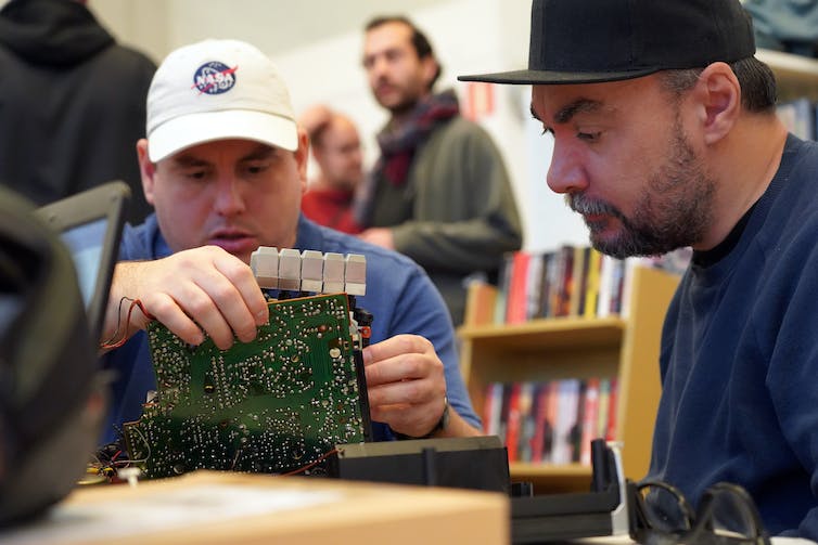 in a library, a man in a white cap holds a circuit board while another man walks past