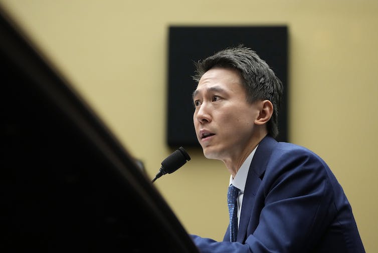 An Asian man in a navy suit and tie speaks into a microphone from behind a desk.