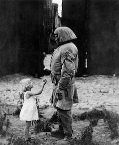 A black and white photo of a little girl in a white dress holding up a piece of fruit to a huge man in dirty clothes in an alleyway.