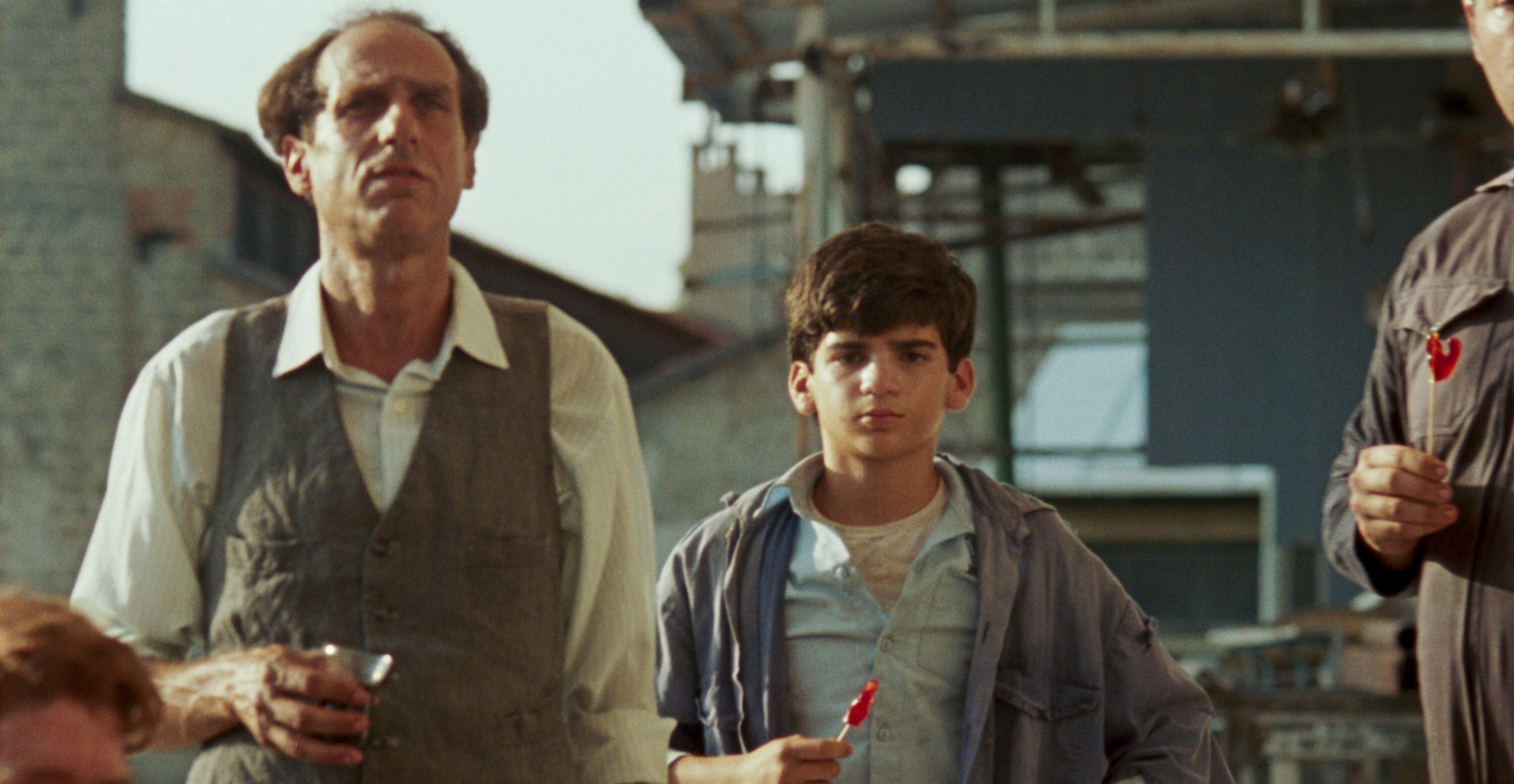 A young boy eating a lollipop staring forward while standing next to a balding man holding a glass in his hand in June Zero.